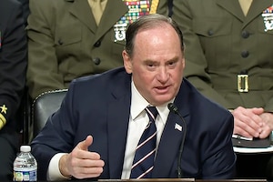 A man in business attire sits at a table while speaking into a microphone. Behind him are three seated uniformed military personnel.