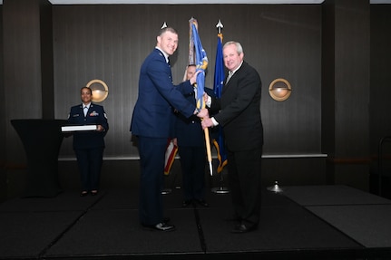 Maj. Aleksander Layne, 433rd Logistics Readiness Squadron commander, hands the 433rd Airlift Wing guidon to Jay Gilmore, SWBC Mortgage Company senior loan officer, during the 433rd AW 2025 Honorary Commanders’ Induction Ceremony in San Antonio, June 7, 2025.