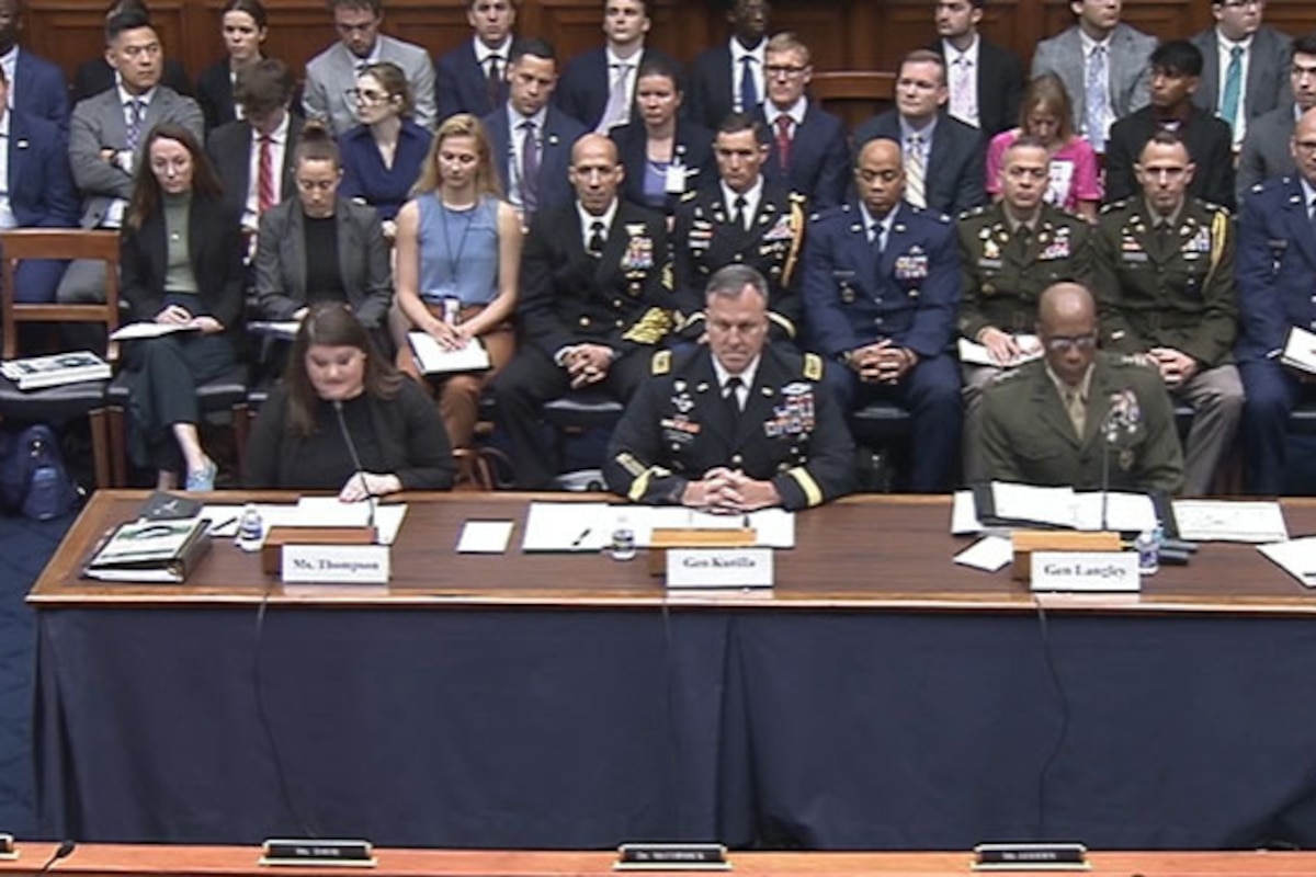 A woman wearing business attire sits at a desk and speaks into a microphone; two men wearing military uniforms are seated next to her.