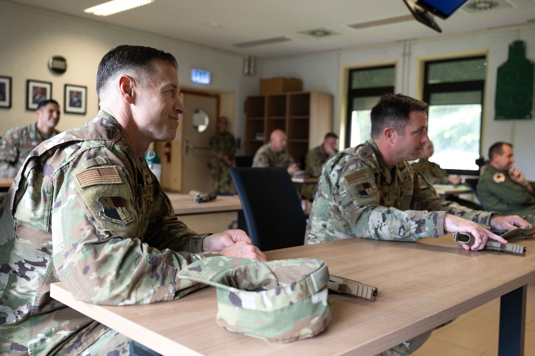 Two Airmen sit at a desk and listen to instruction about handgun firing procedures.