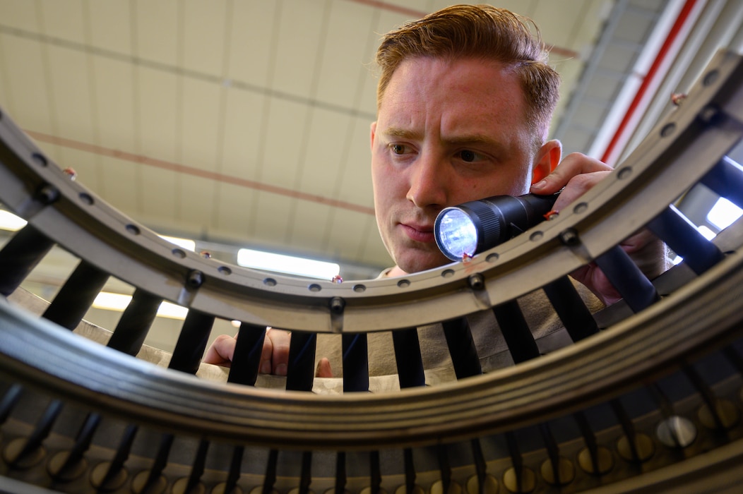 An Airman uses a flashlight to closely inspect components inside a jet engine.