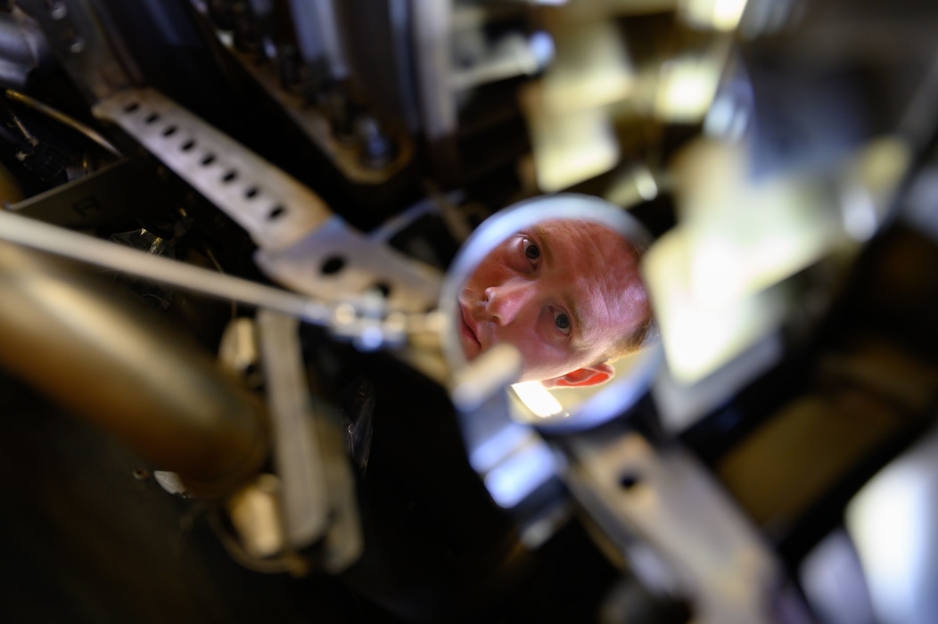 An Airman is visible in the reflection of a mirror as he uses it to inspect the internal parts of a jet engine.