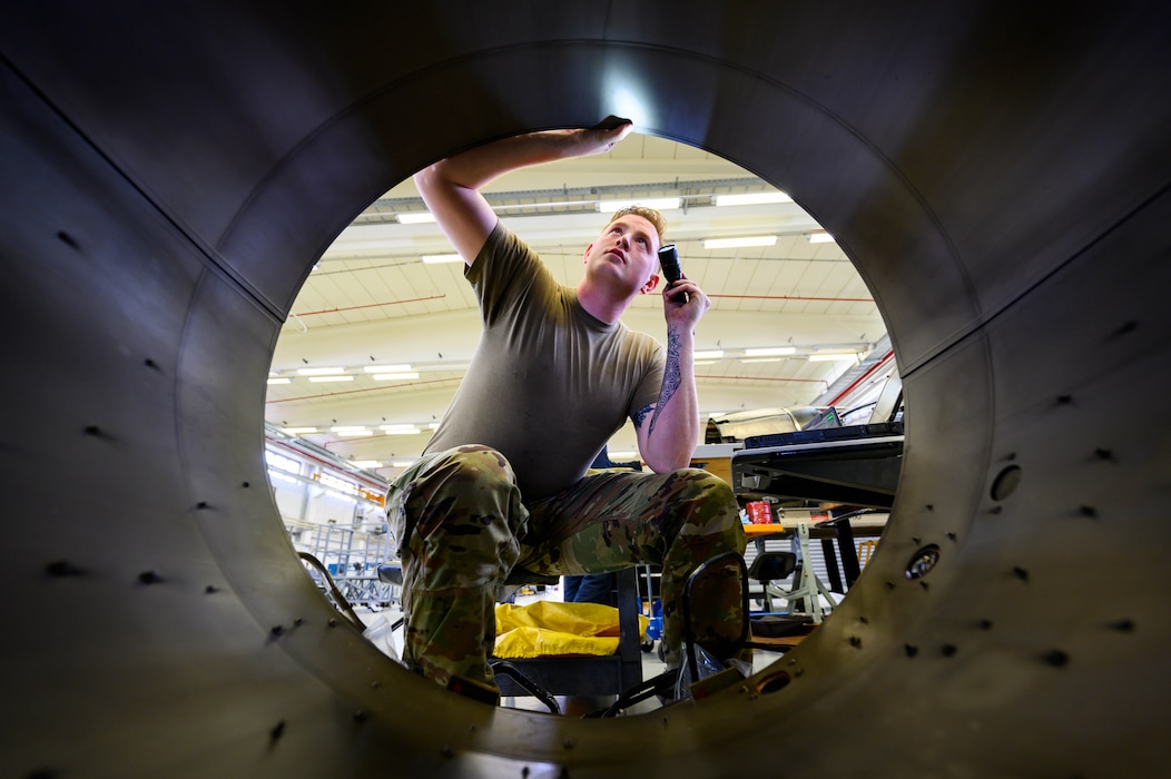 An Airman inspects a large, tunnel-like engine component with a light.