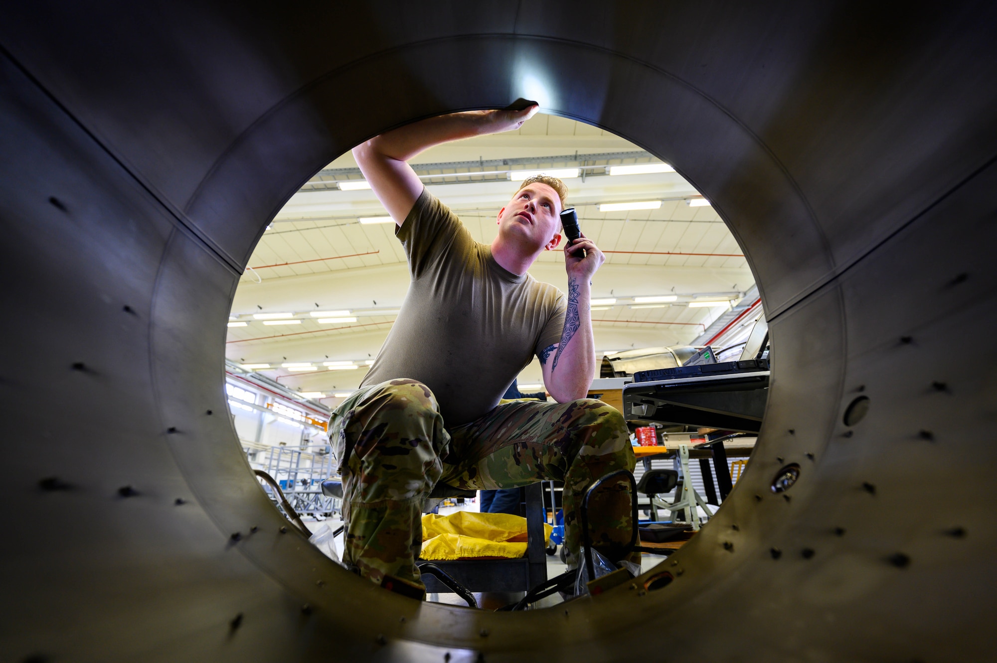 An Airman inspects a large, tunnel-like engine component with a light.