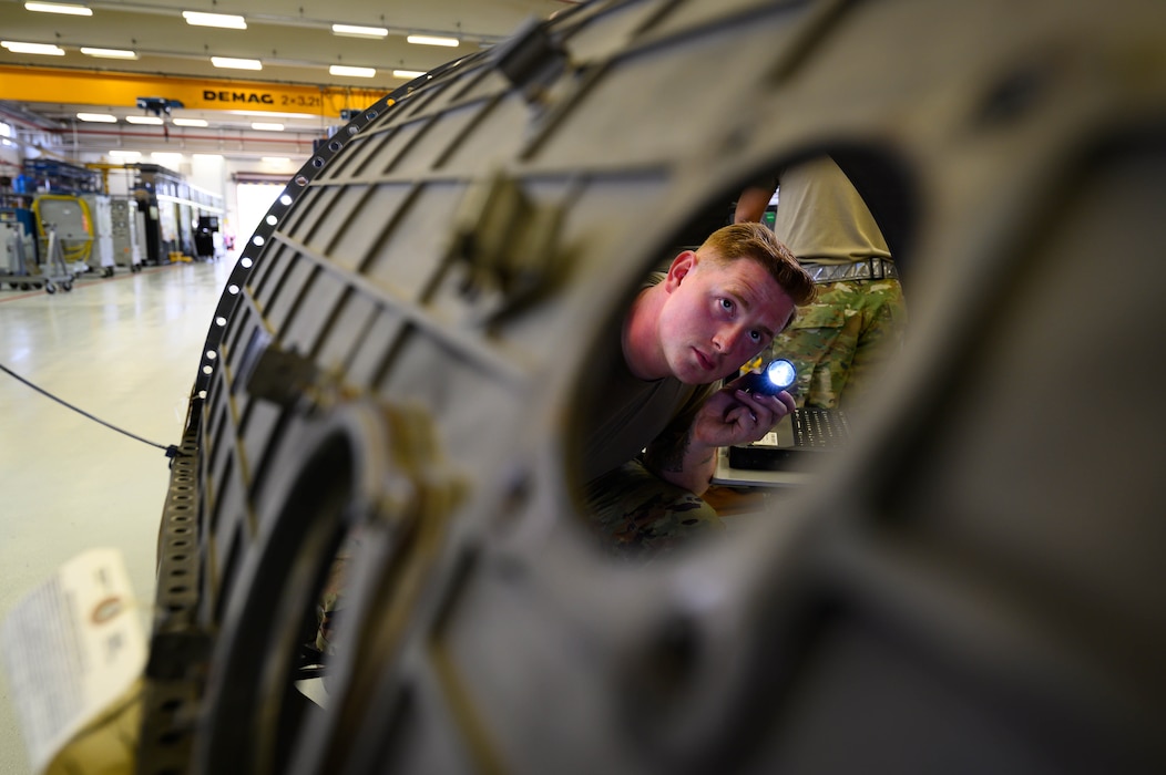An Airman inspecting a jet engine with a flashlight is visible through a hole in one of the parts.