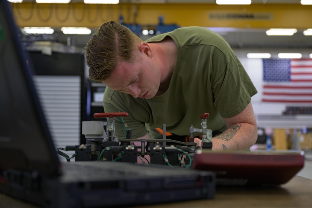 An Airman is leaning over a desk, working on a jet engine component.