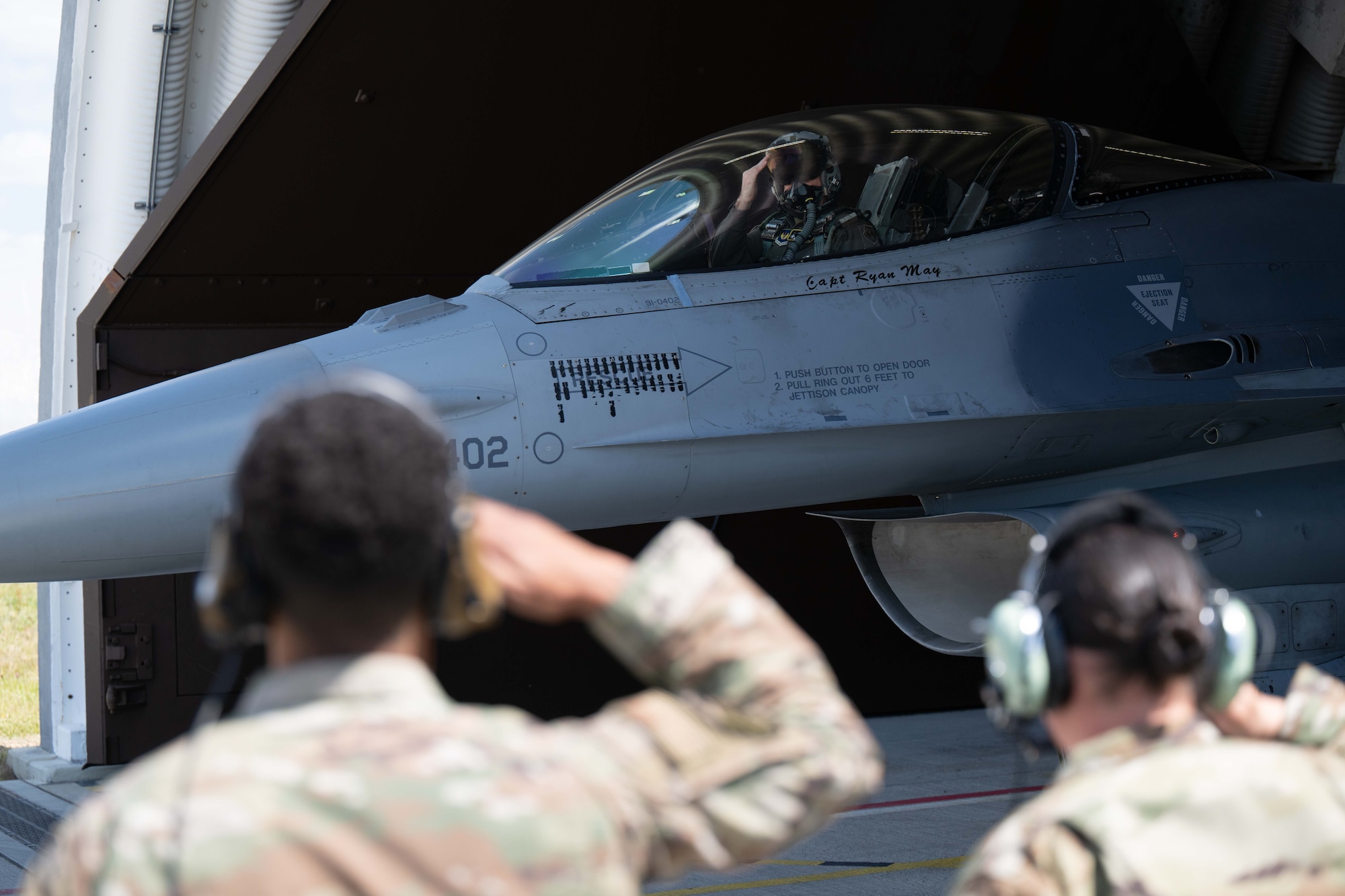 Two Airmen salute a pilot who is taxiing out of a hardened aircraft shelter in his fighter aircraft.