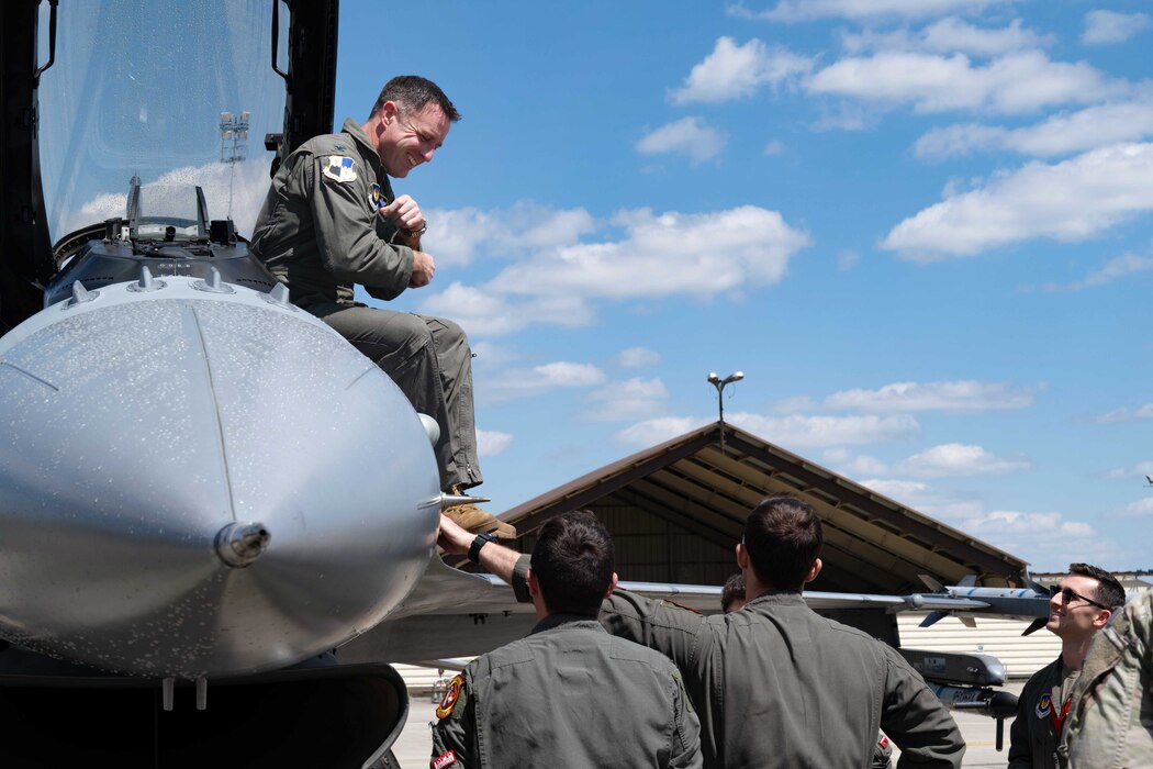 A pilot sits on the side of his fighter aircraft's canopy, with other pilots smiling up at him from the ground.