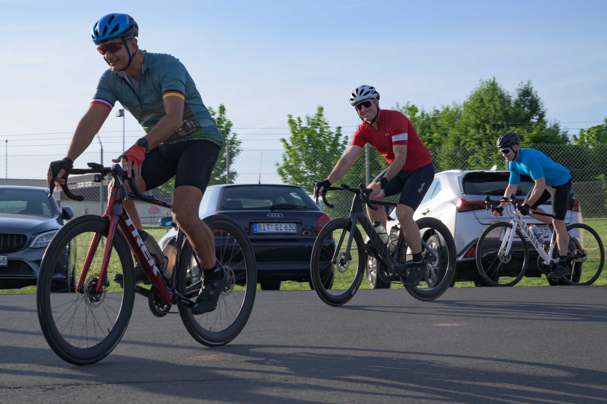 Three cyclists depart from a visitor's center parking lot on bicycles.