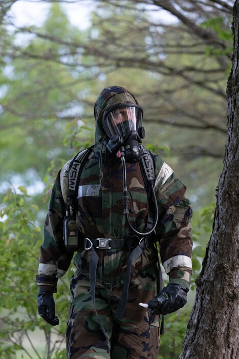 An Airman in full protective equipment walks through a grove of trees.