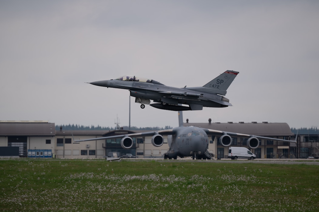 A fighter aircraft takes off from a runway with a larger cargo aircraft in the background.