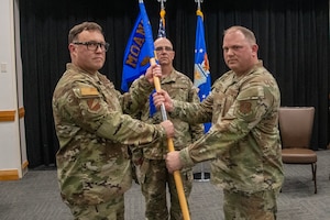 Two U.S. Air National Guard Airmen pose for a photo while handing off a guidon flag. They are wearing OCP pattern uniforms.