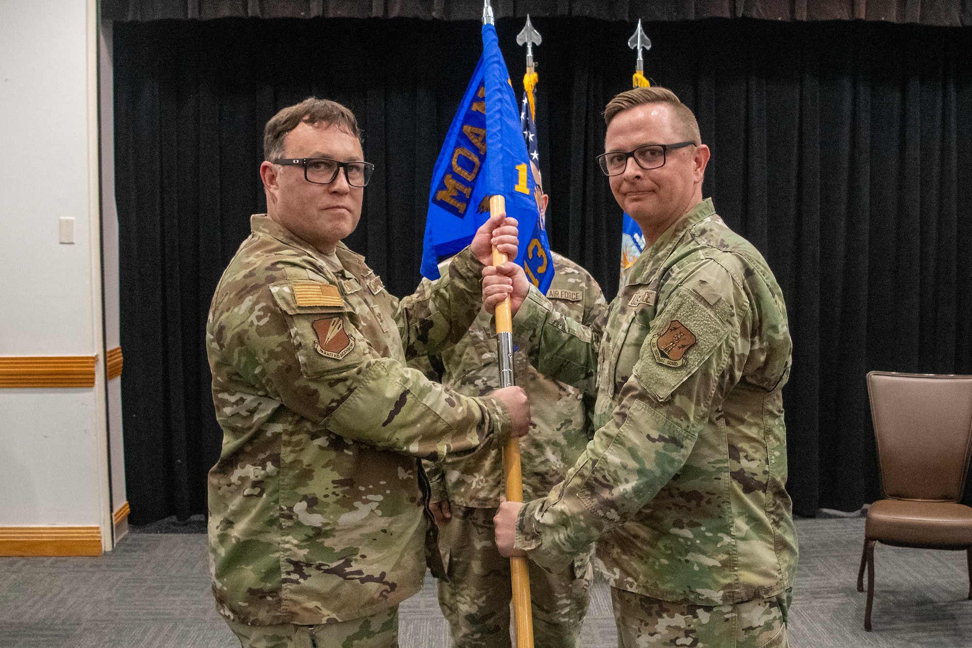 Two U.S. Air National Guard Airmen pose for a photo while handing off a guidon flag. They are wearing OCP pattern uniforms.