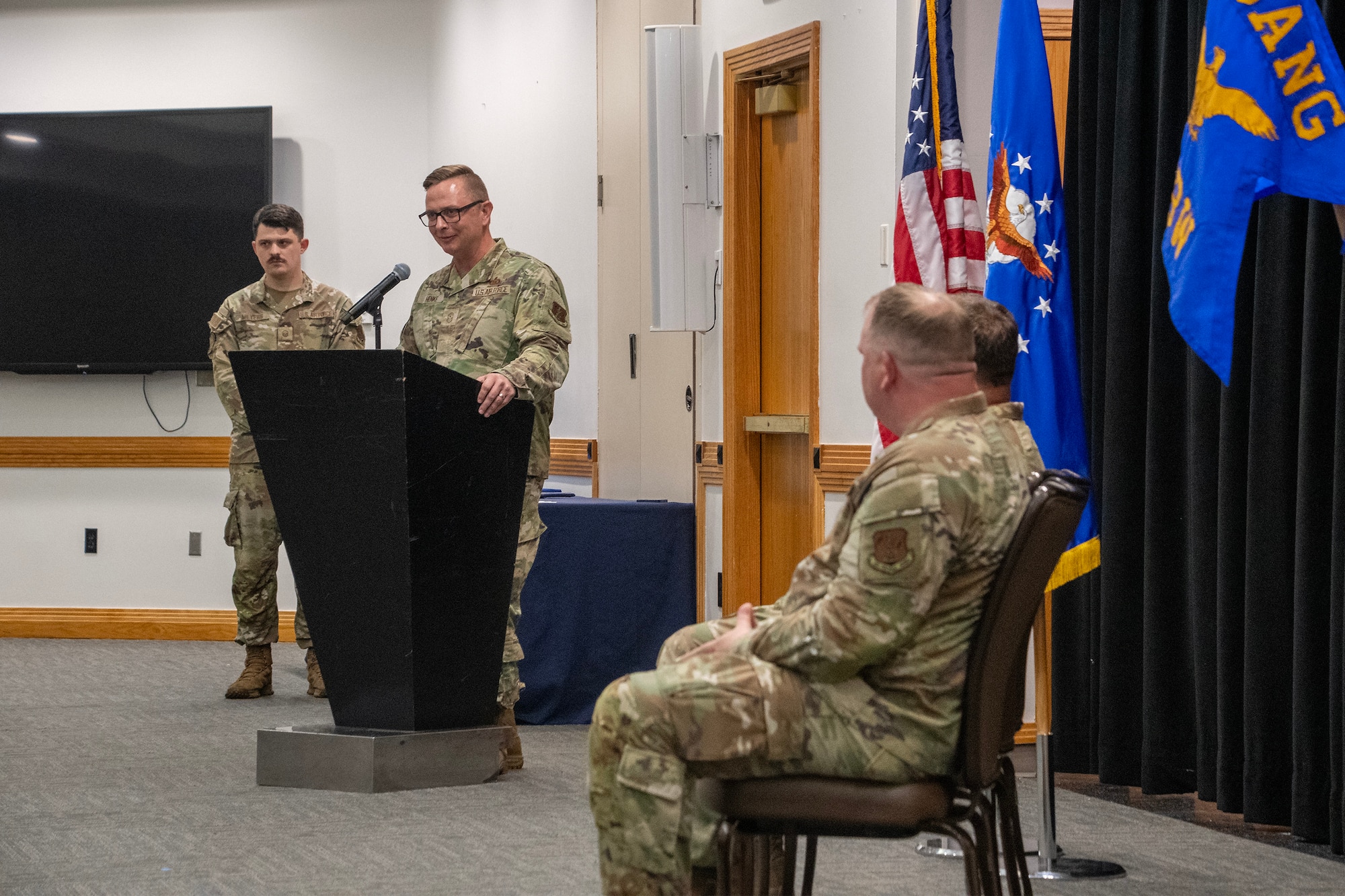 A U.S. Air National Guard Airman stands at a lectern giving a speech during a ceremony. Two Airmen are seated before U.S. and Air Force flags to his left. They are all wearing OCP pattern uniforms.
