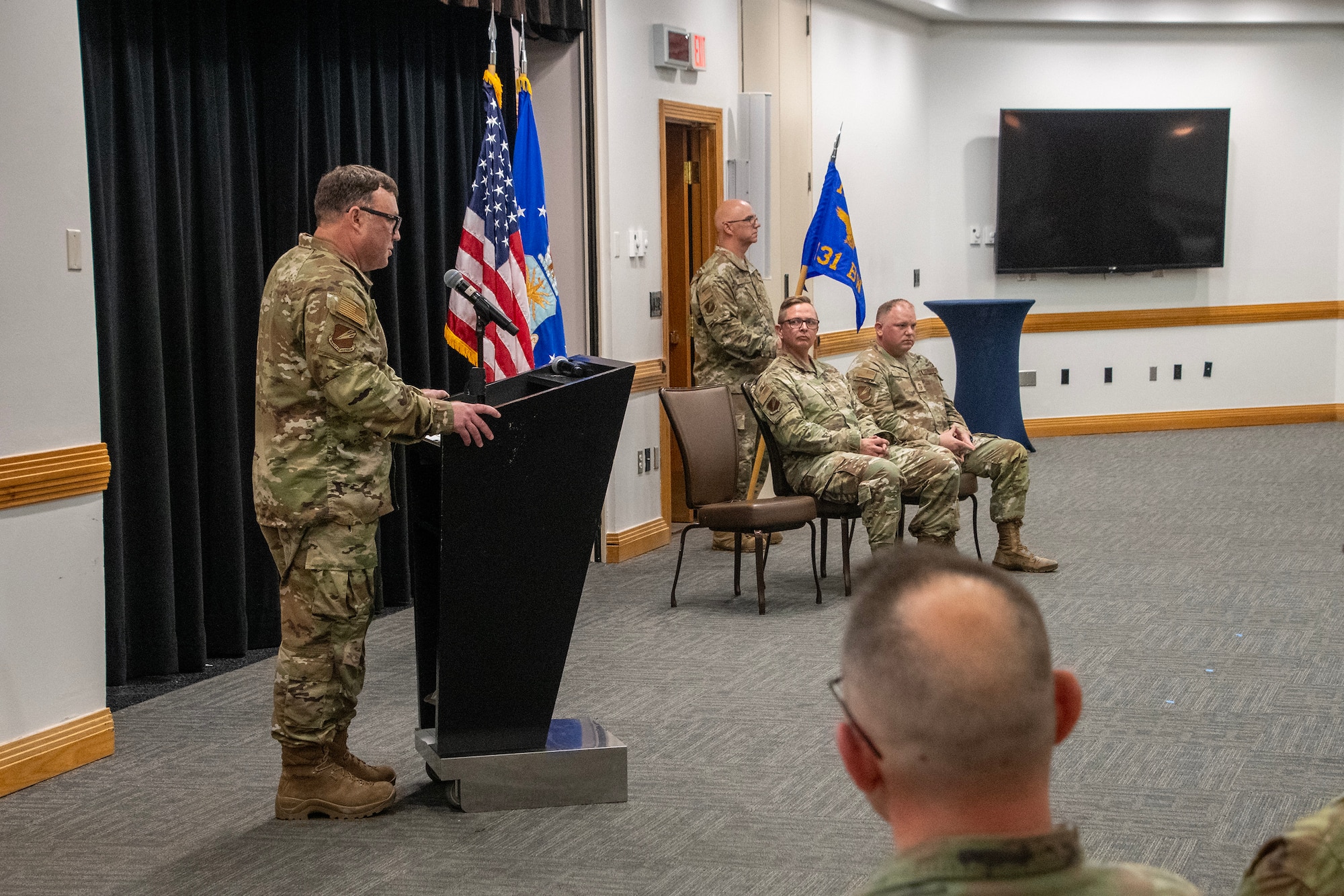 A U.S. Air National Guard Airman stands at a lectern giving a speech during a ceremony. Two Airmen are seated before U.S. and Air Force flags to his left. They are all wearing OCP pattern uniforms.