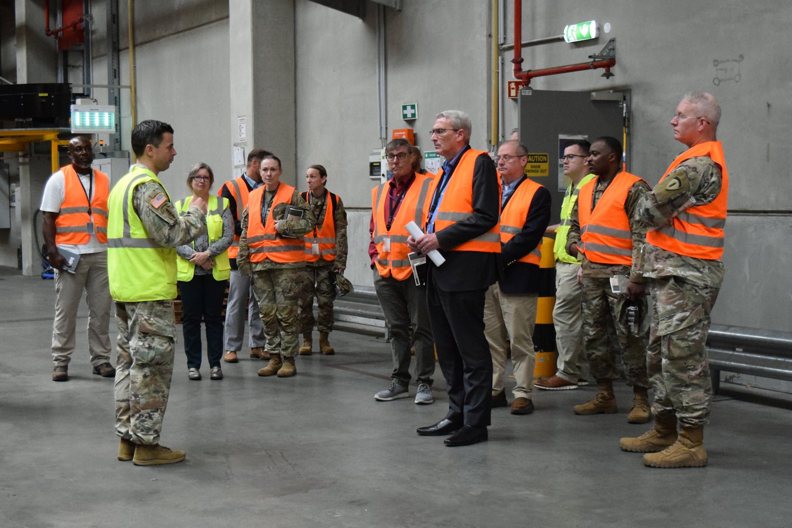 A group of people wearing reflective safety vests stand inside of a warehouse talking to each other.