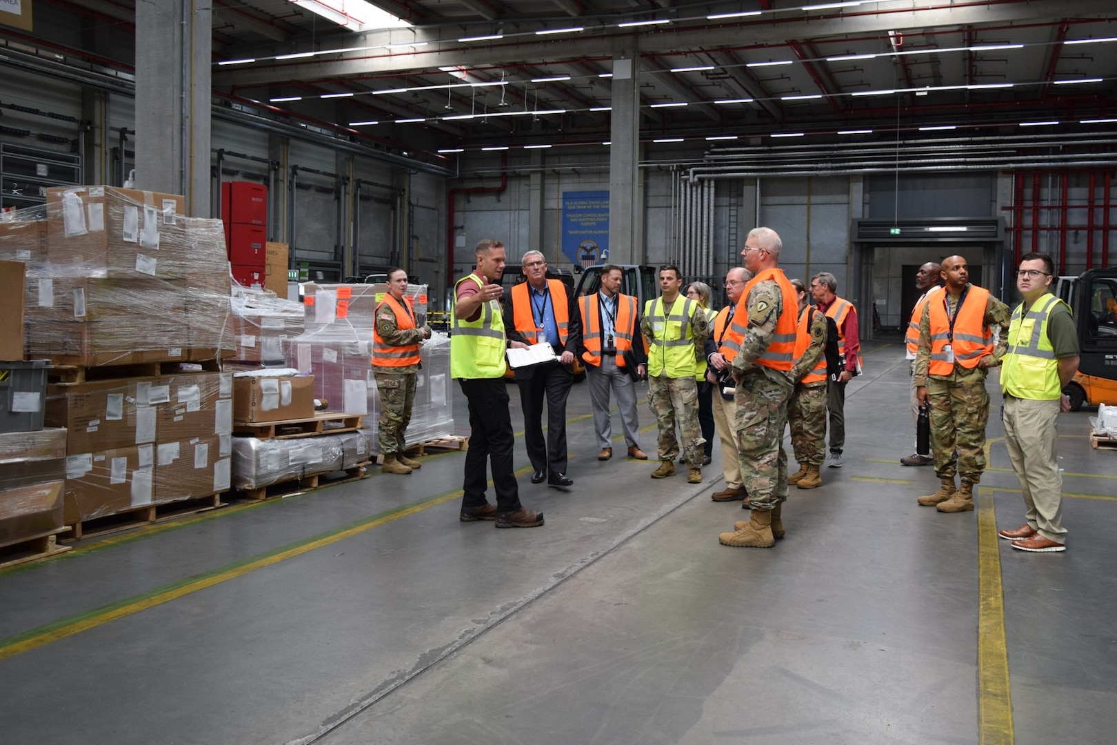 A group of people wearing reflective safety vests stand inside of a warehouse talking to each other.
