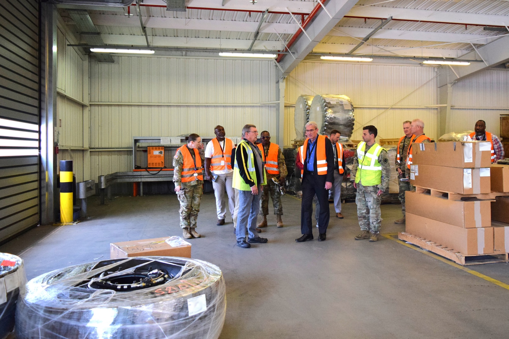 A group of people wearing reflective safety vests stand inside of a warehouse talking to each other.