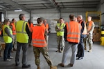 A group of people wearing reflective safety vests stand inside of a warehouse talking to each other.