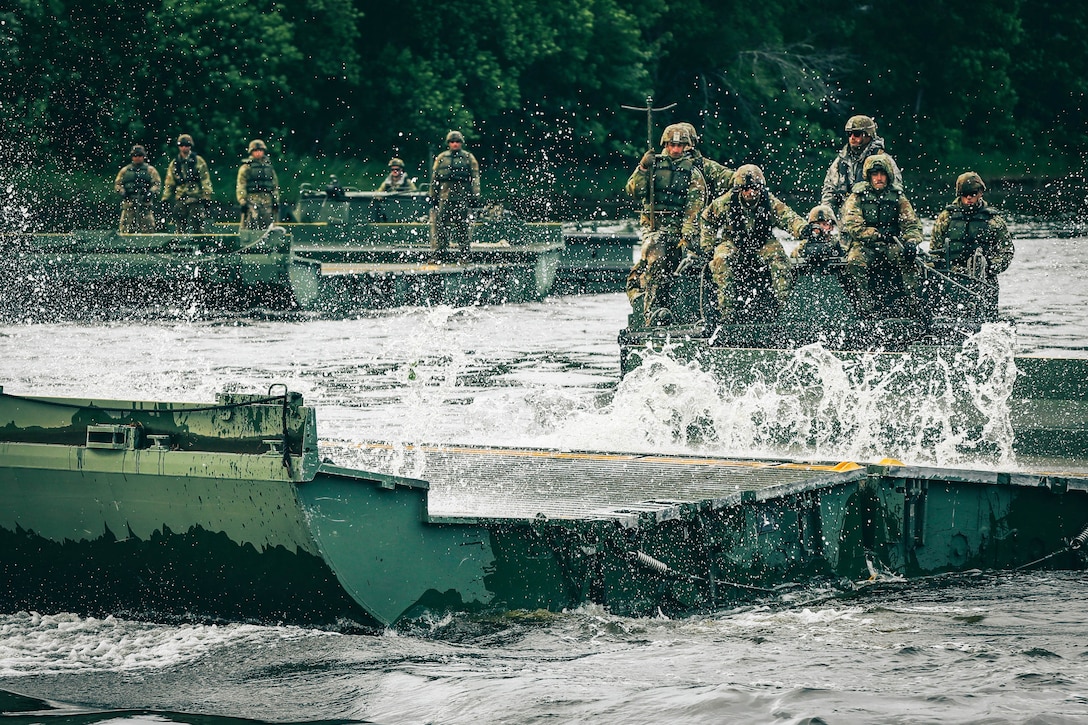 Water splashes as five soldiers in tactical gear ride a raft in a river as fellow soldiers watch near a wooded area.