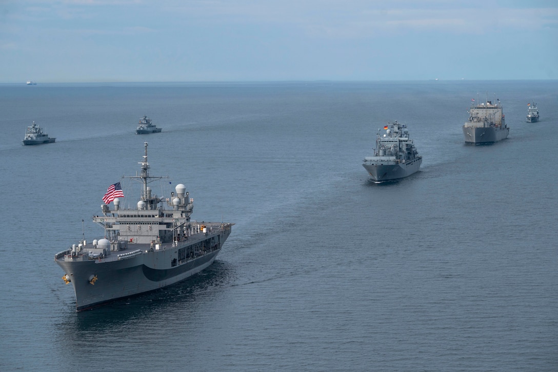 Six ships sail in formation in dark blue water under a blue sky.