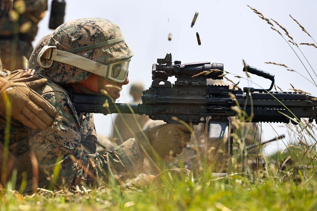 Ammo flies in the air as a Marine wearing protective gear fires a weapon while lying in tall grass against a blue sky.