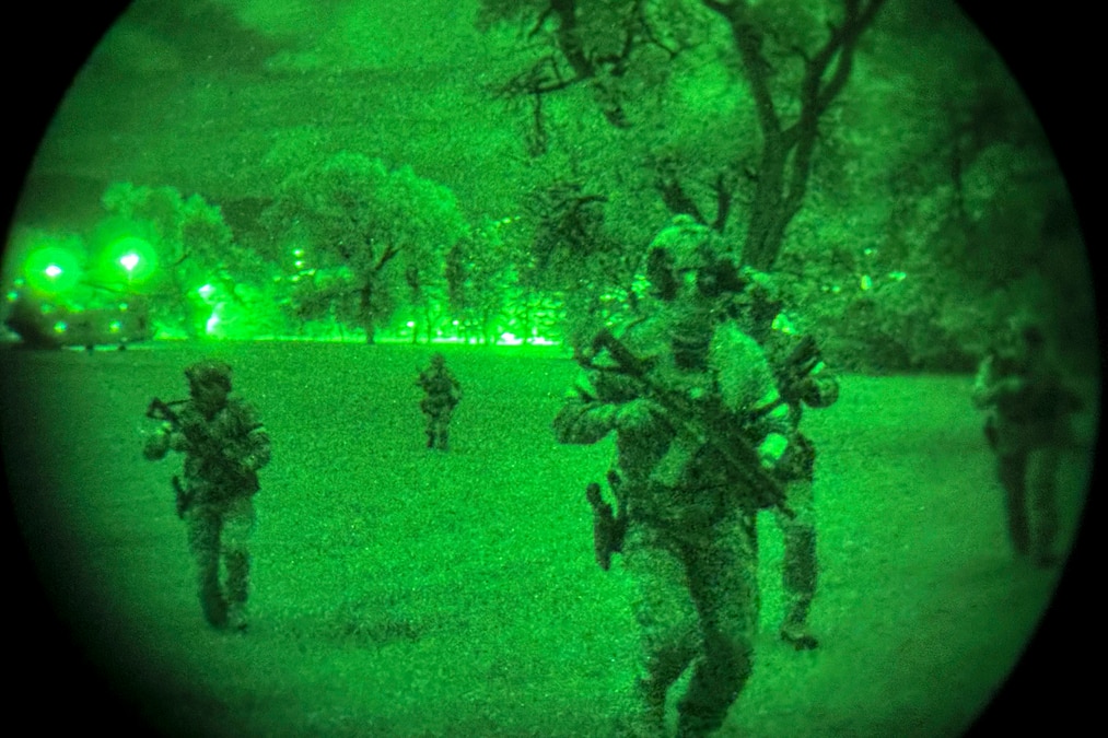 U.S. Army, British and Polish troops in tactical gear run carrying weapons run from a distant helicopter near trees as seen through night vision lens.