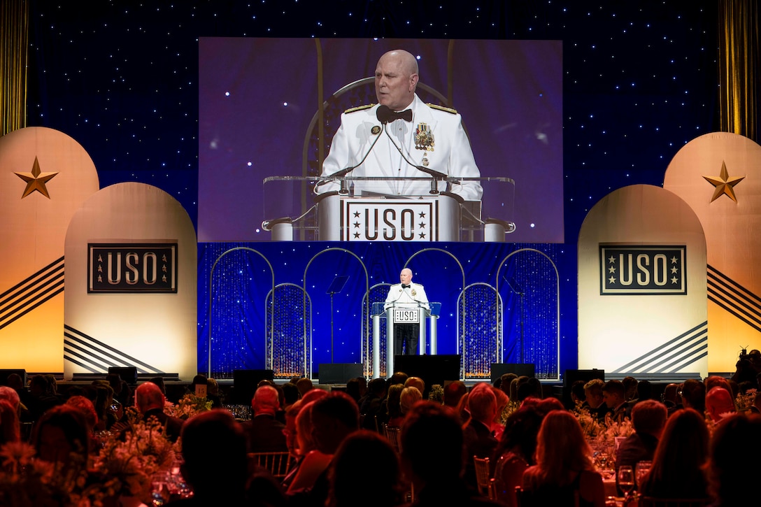 A man in a white suit stands behind a lectern and speaks to an audience sitting at tables. Behind him, a large screen shows a close-up of the man at the lectern.