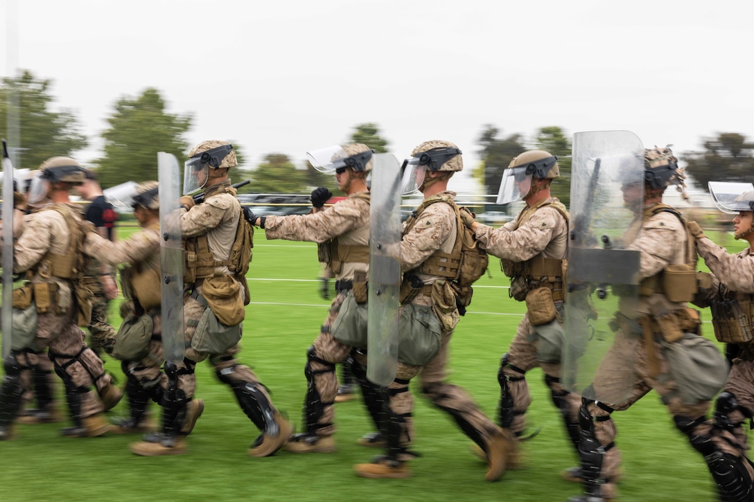 Marines in combat gear and holding plastic shields run in formation on a grassy field.
