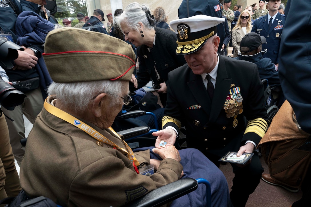 A man in a black military suit and white hat knees to give a pin to a veteran wearing a military outfit.