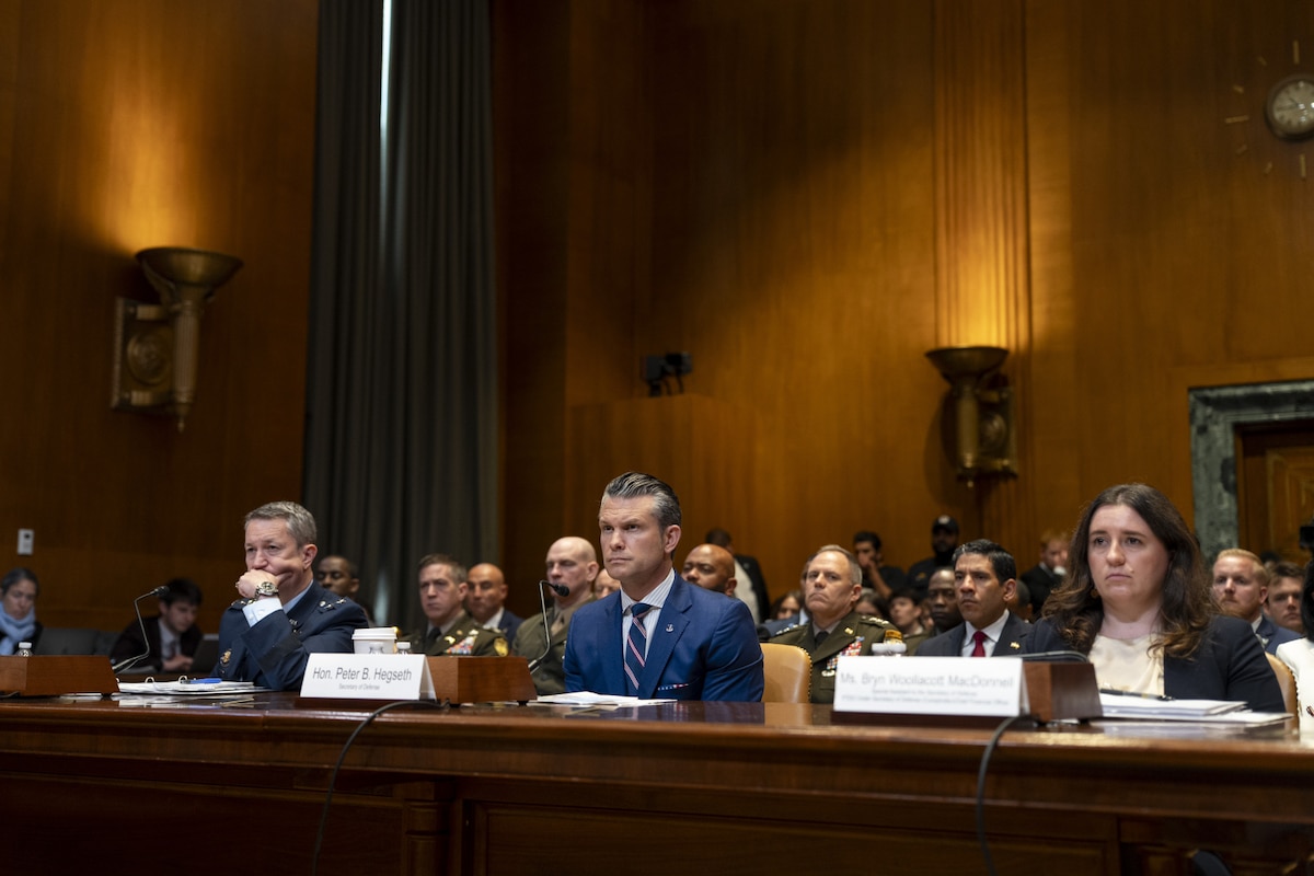 Three individuals sit at a long wooden table inside a large room with wooden paneling. Two are wearing business attire, and one is wearing a dress military uniform. People are sitting behind them.