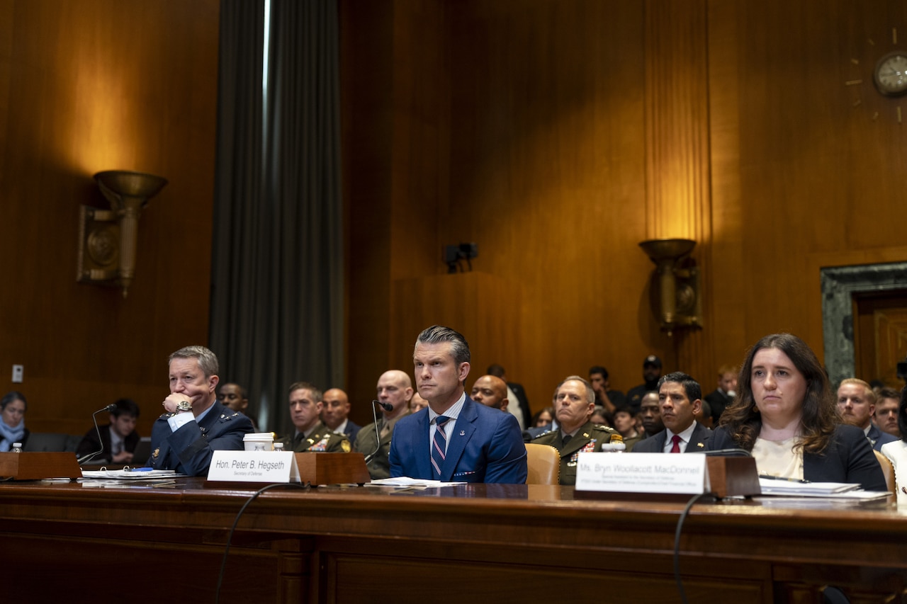 Three individuals sit at a long wooden table inside a large room with wooden paneling. Two are wearing business attire, and one is wearing a dress military uniform. People are sitting behind them.