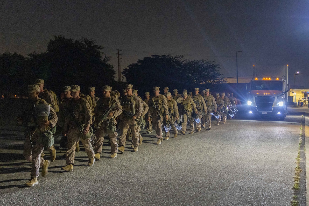 Marines walk in a row on a paved surface with a dark sky and trees in the background.