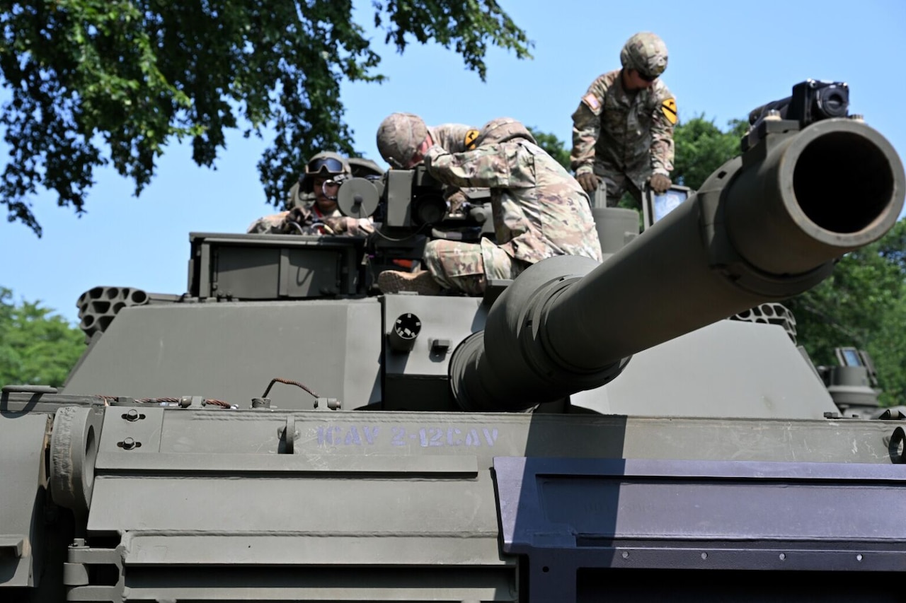 People in camouflage military uniforms are sitting atop a military tank outdoors.