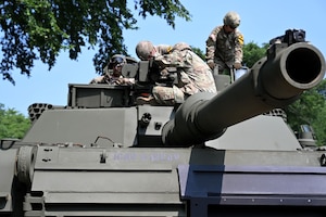People in camouflage military uniforms are sitting atop a military tank outdoors.