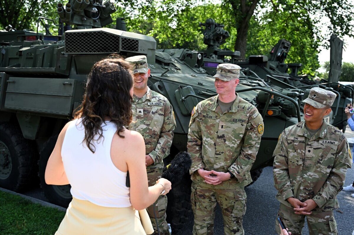 Three men in camouflage military uniforms are standing in front of a large military vehicle while speaking to a woman in civilian attire with her back to the camera.