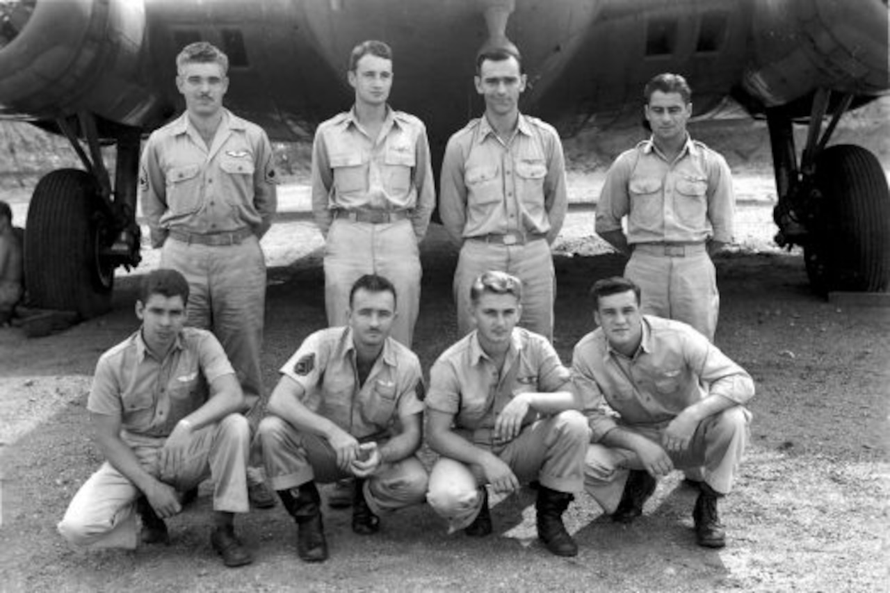 Four men in military uniforms squat down, and four stand to pose for a photo in front of an aircraft.
