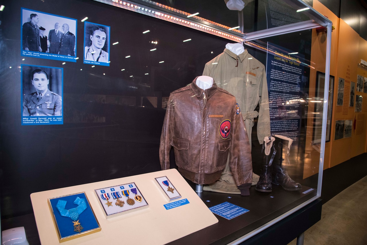 Photos, medals, two jackets and a pair of boots are shown in a display case against a wall.