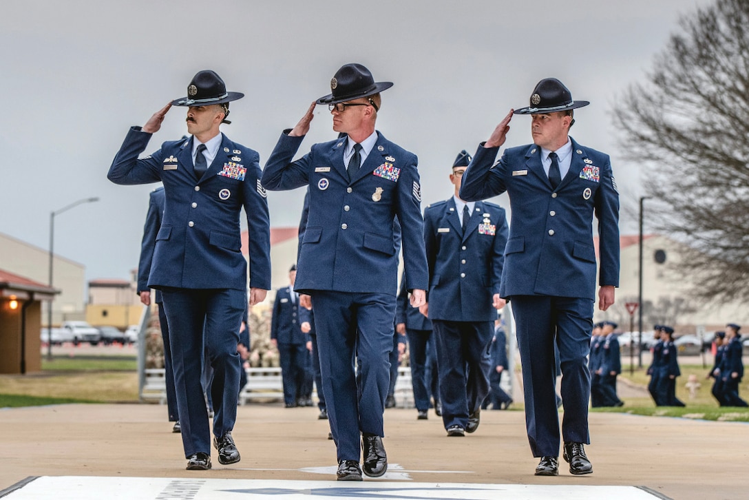 Three U.S. Air Force military training instructors in dress blue uniforms and campaign hats march forward while saluting. They are followed by other uniformed graduates in formation.