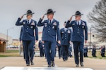 Three U.S. Air Force military training instructors in dress blue uniforms and campaign hats march forward while saluting. They are followed by other uniformed graduates in formation.