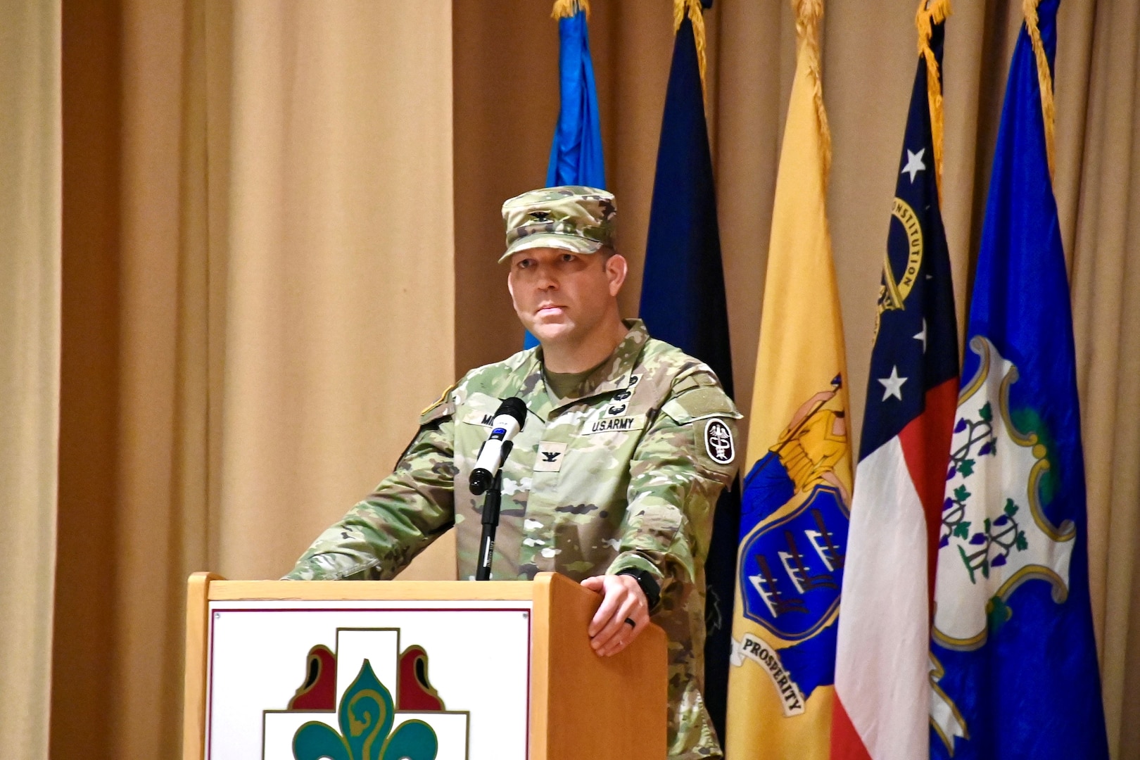 Col. Miller speaks behind a podium with the BJACH Crest attached to the front.