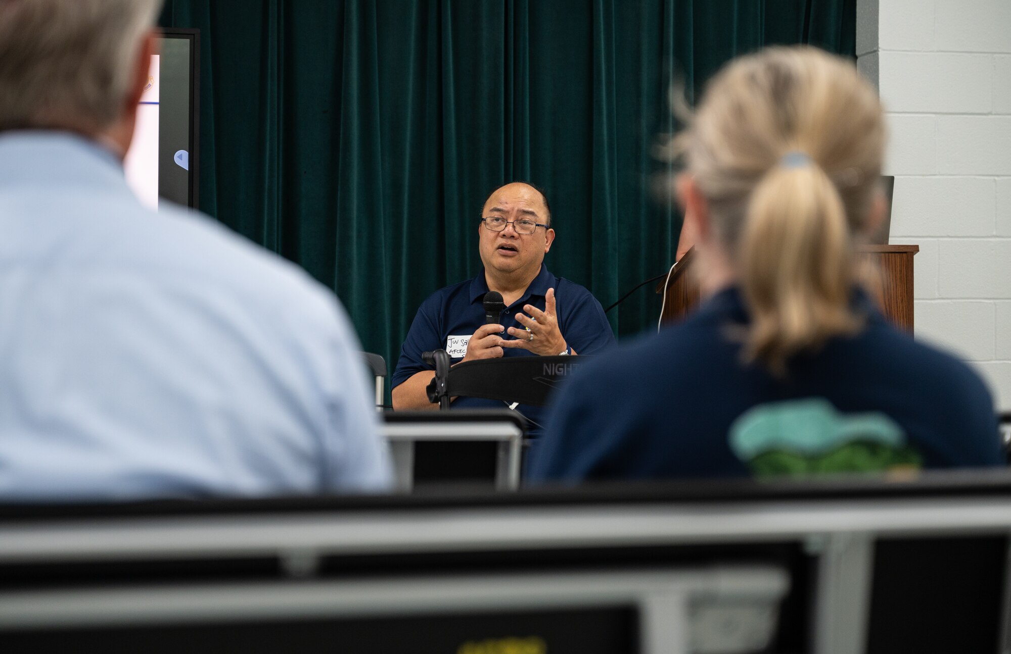 A man sits and speaks into a microphone as people listen.
