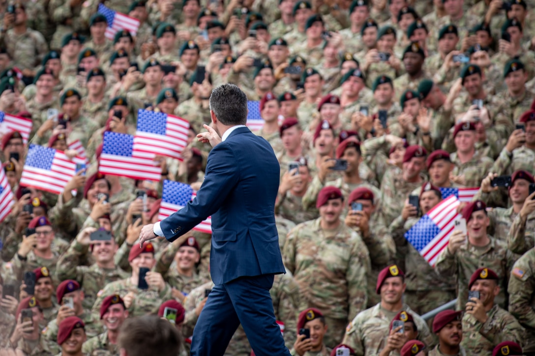 A man in a suit walks in front of a crowd of soldiers. The man is pointing at the crowd.