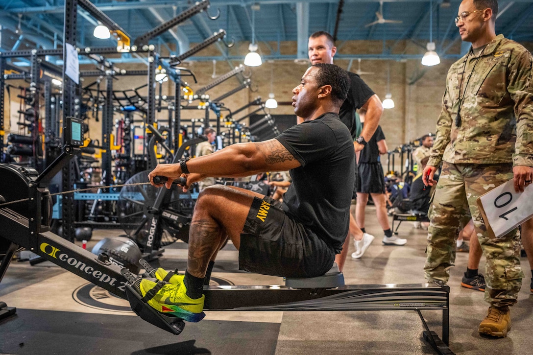 A soldier uses a rowing machine in an indoor gym as other soldiers stand and watch.