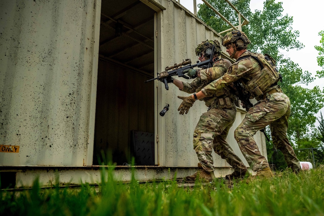 Two airmen in combat gear, one aiming a rifle and one tossing a grenade-like device into an opening of a metal container outdoors, prepare to enter the container.