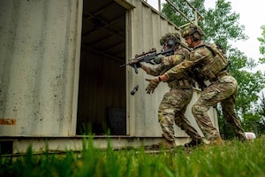 Two airmen in combat gear, one aiming a rifle and one tossing a grenade-like device into an opening of a metal container outdoors, prepare to enter the container.
