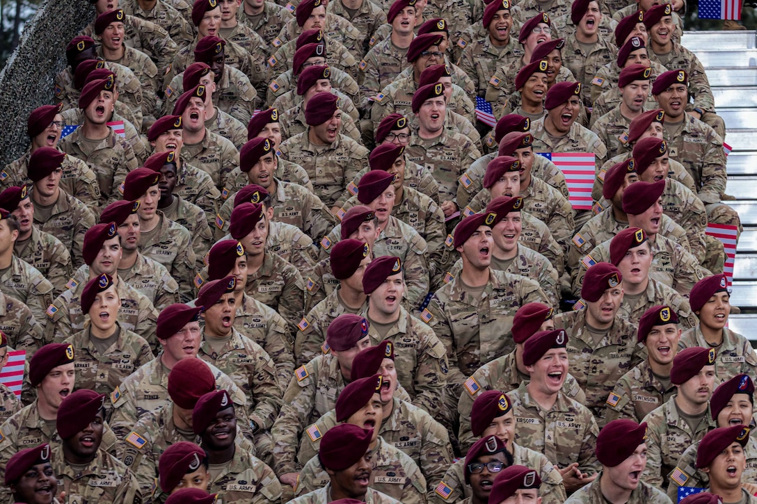 A group of soldiers wearing maroon berets and sitting in an outdoor bleacher yell in unison.