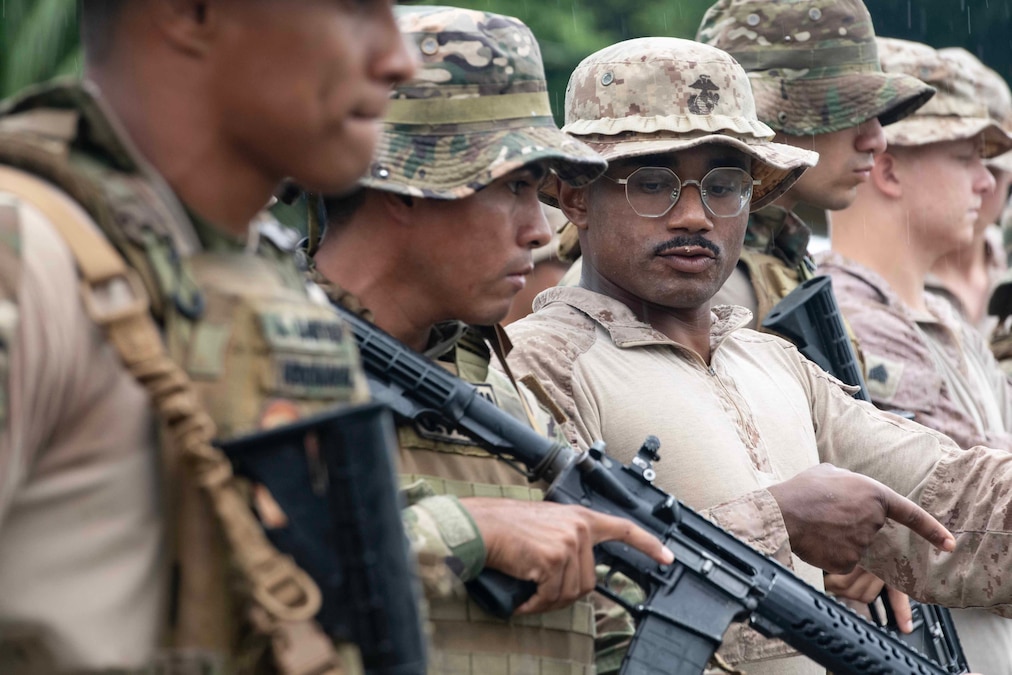 A Marine talks to a member of the Panamanian military holding a rifle outdoors.