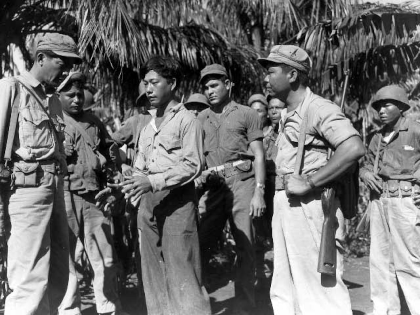 A black and white photo with a group of men standing in front of palm trees.