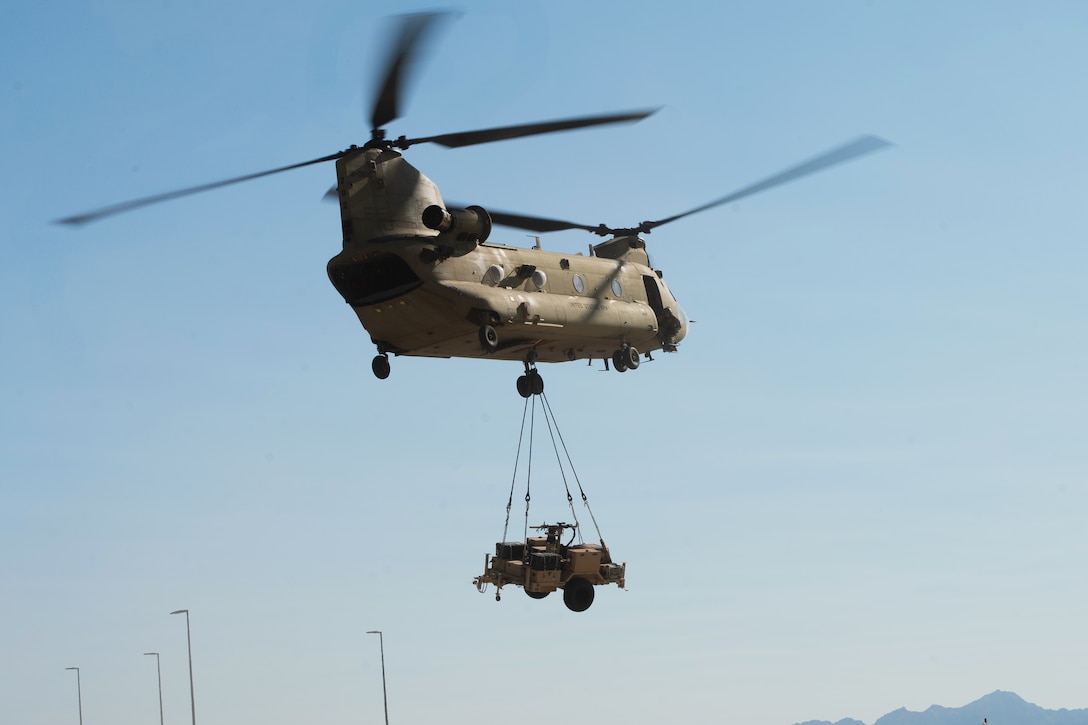 A flying helicopter carries a heavy piece of equipment attached by four chains to the bottom of the aircraft on a cloudless day.